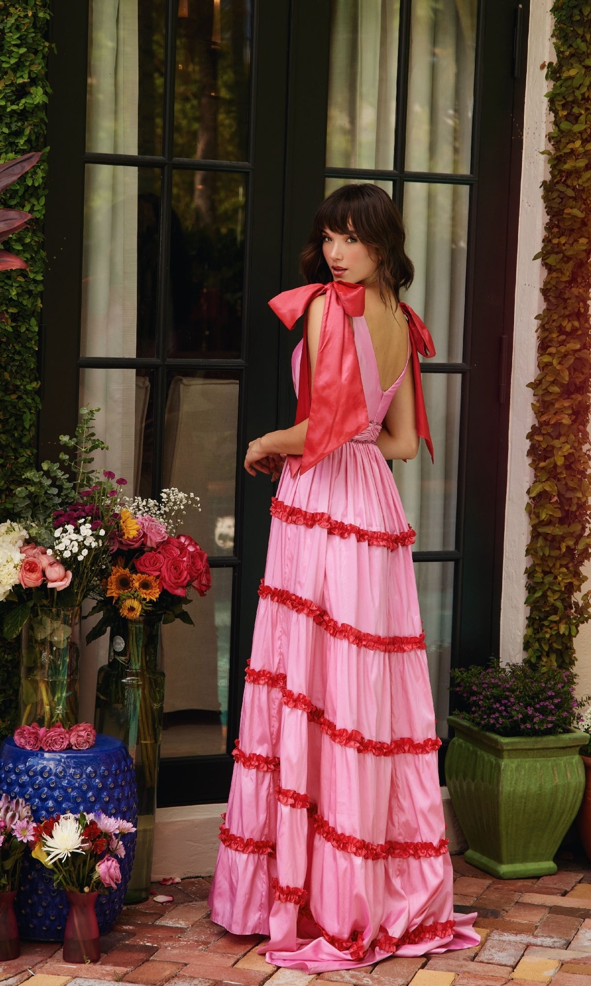 A woman wearing the Shoulder Bows A-Line Formal Dress: Ava Presley 40091 stands on a brick patio before a glass door, surrounded by colorful flower arrangements in assorted vases.